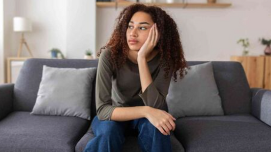“Young woman sitting on a sofa with her head resting on her hand, looking thoughtful and emotionally drained.”
