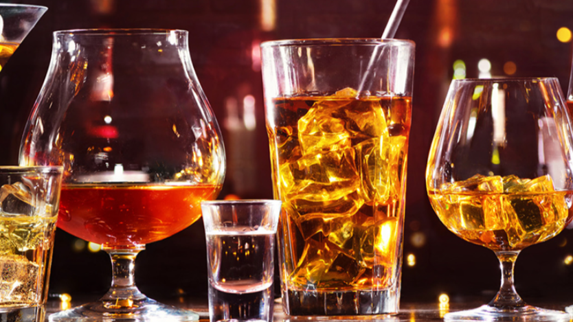 A selection of amber-coloured alcoholic drinks in different glasses, filled with ice and glowing in warm bar lighting, symbolising how alcohol shows up in mid-life health and energy.